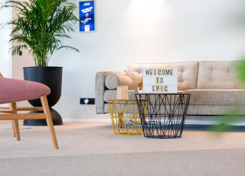 Modern office lounge with pink chair, grey sofa, wire tables, a 'Welcome to Epic' lightbox sign, and a wall-mounted Joan display.
