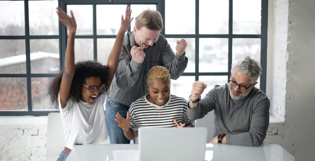 Four colleagues celebrating enthusiastically around a laptop in a bright modern office.