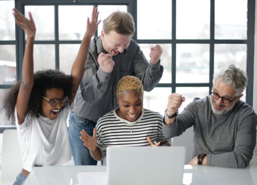 Four colleagues celebrating enthusiastically around a laptop in a bright modern office.