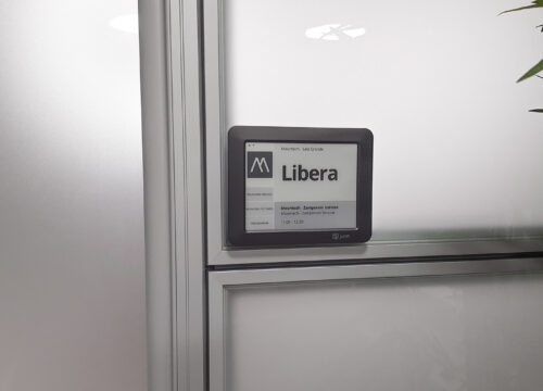 Joan 6 e-ink display mounted at meeting room entrance showing room 'Libera' as available with upcoming schedule.