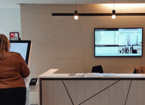 Woman at a visitor kiosk in a modern office lobby with a digital signage display showing room schedules on the wall.