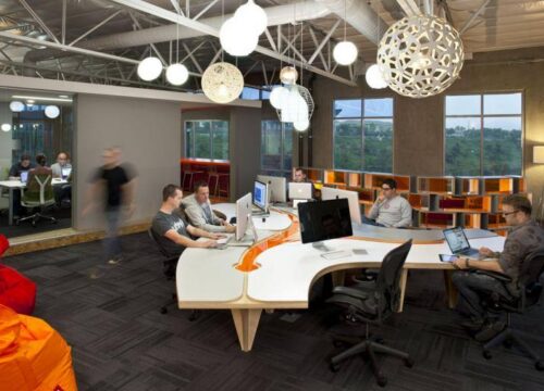 Employees working at a modern open-plan office with white curved desks, globe pendant lights, and large windows.