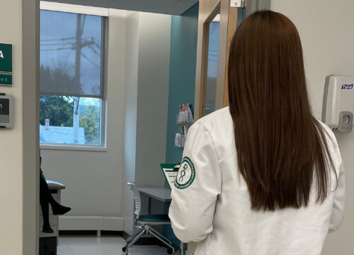 Pharmacy student in white lab coat approaching a simulation room with a Joan e-ink display mounted beside the door.