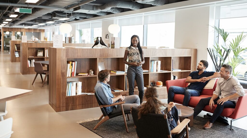 Four colleagues having an informal discussion in a modern open-plan office lounge with warm wood shelving.