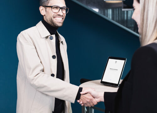 Smiling man shaking hands with a receptionist at a modern office, visitor welcome screen visible on a nearby tablet.