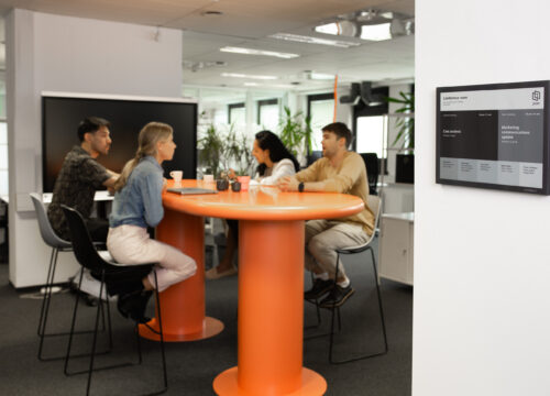 Joan 13 e-ink display mounted on office wall showing conference room schedule, with four colleagues meeting at an orange table.