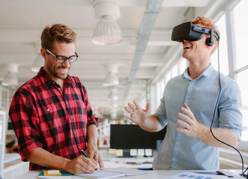 Two professionals in a modern office explore VR technology, one wearing a headset laughing, the other taking notes.