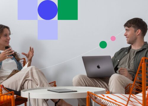 Two colleagues having a conversation in a modern office lounge, one with a MacBook, surrounded by colorful graphic accents.
