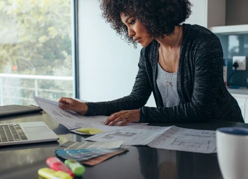 Woman reviewing floor plan documents at a home office desk with a laptop, color swatches, and coffee mug nearby.