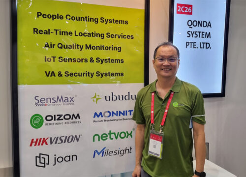 Exhibitor standing in front of a trade show booth banner listing IoT and workplace tech partners, including Joan.