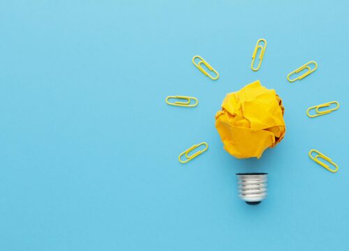 Crumpled yellow paper ball on a light bulb base, surrounded by yellow paper clips as rays, symbolizing innovation.