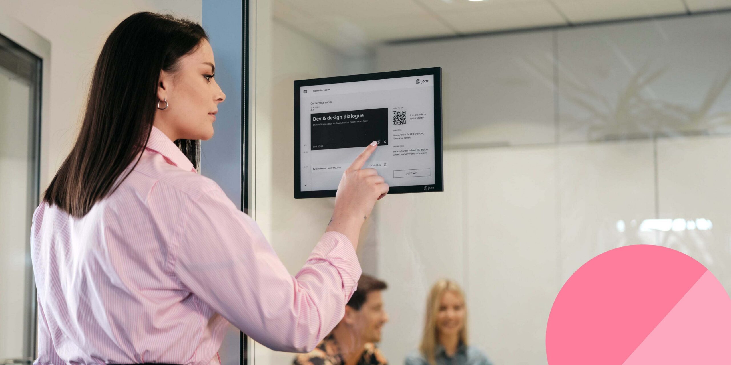 Woman pointing at a Joan 13 e-ink display mounted on a glass wall showing a meeting room schedule.