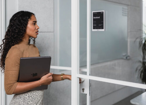 Woman with laptop reaching for a meeting room door, checking a Joan 6 Pro e-ink display showing 'Booked'.