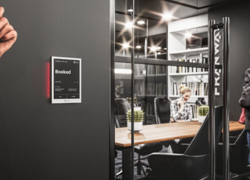 Joan 6 Pro e-ink display mounted outside a glass meeting room showing a 'Booked' status with red LED indicator.