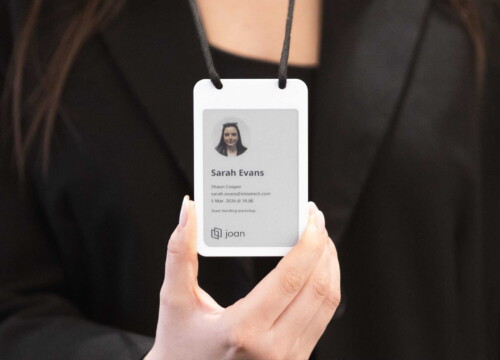 Hand holding a Joan e-ink visitor badge displaying guest name, host, email, and meeting details.
