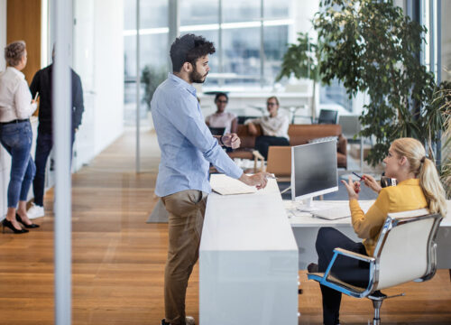 Visitor checking in at a modern office reception desk, greeted by a receptionist in a bright open workspace.