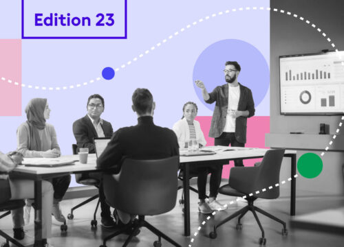 Diverse team in a meeting room watching a presenter gesture toward a data dashboard on a large screen.