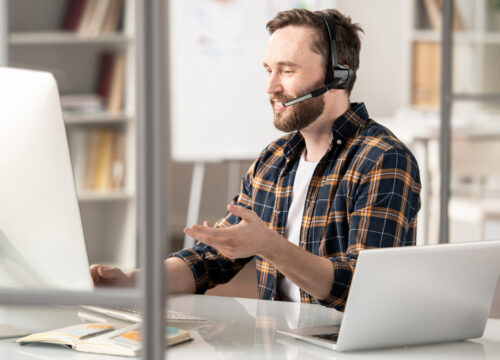 Man in plaid shirt wearing a headset, speaking during a video call at an office desk with a desktop and laptop.