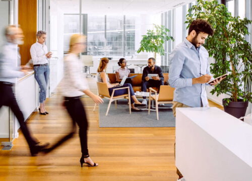 Busy modern office with employees walking, collaborating, and using devices in an open, light-filled workspace.