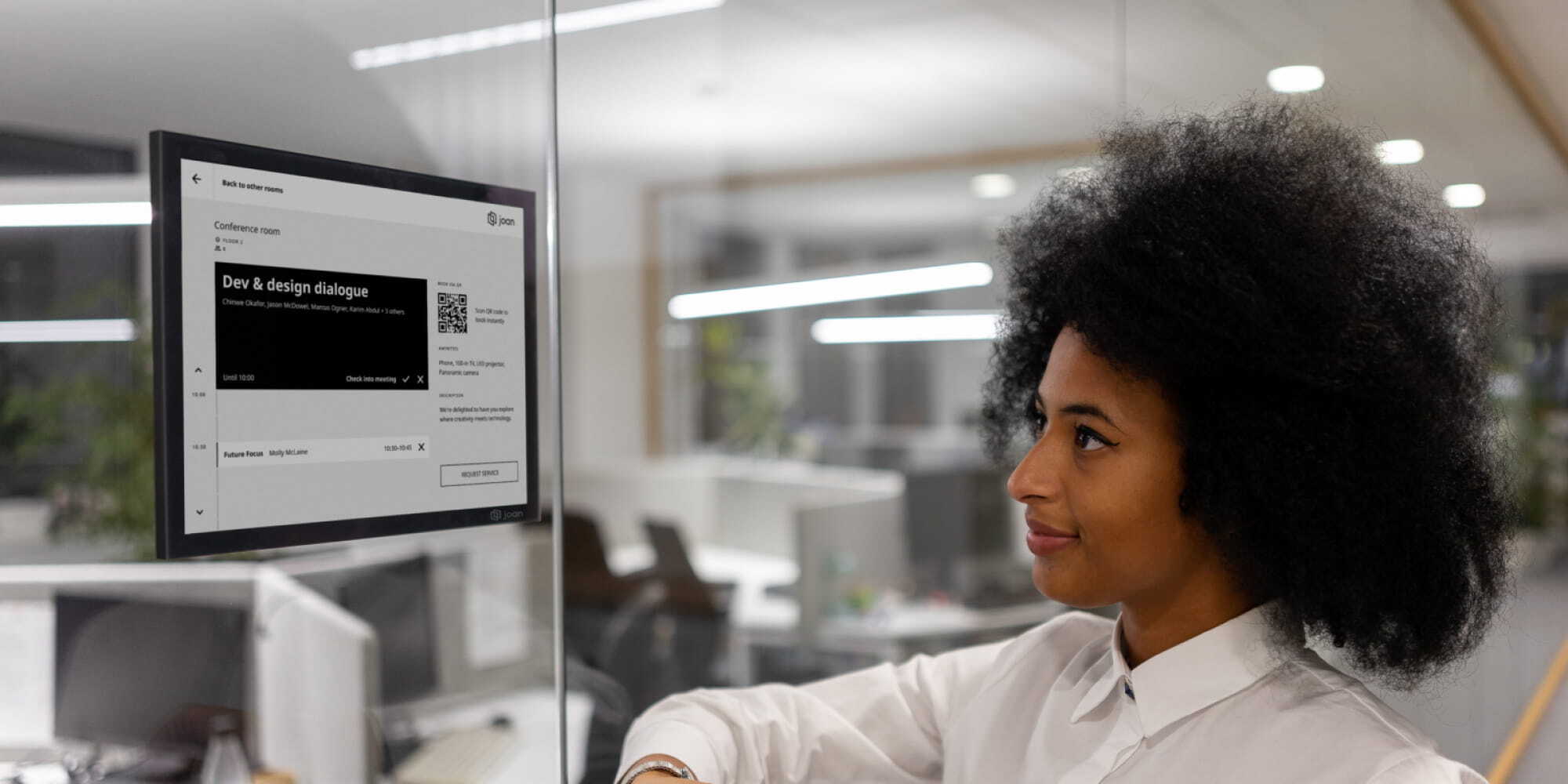 Professional woman viewing a Joan 13 e-ink display showing a conference room schedule on a glass office wall.