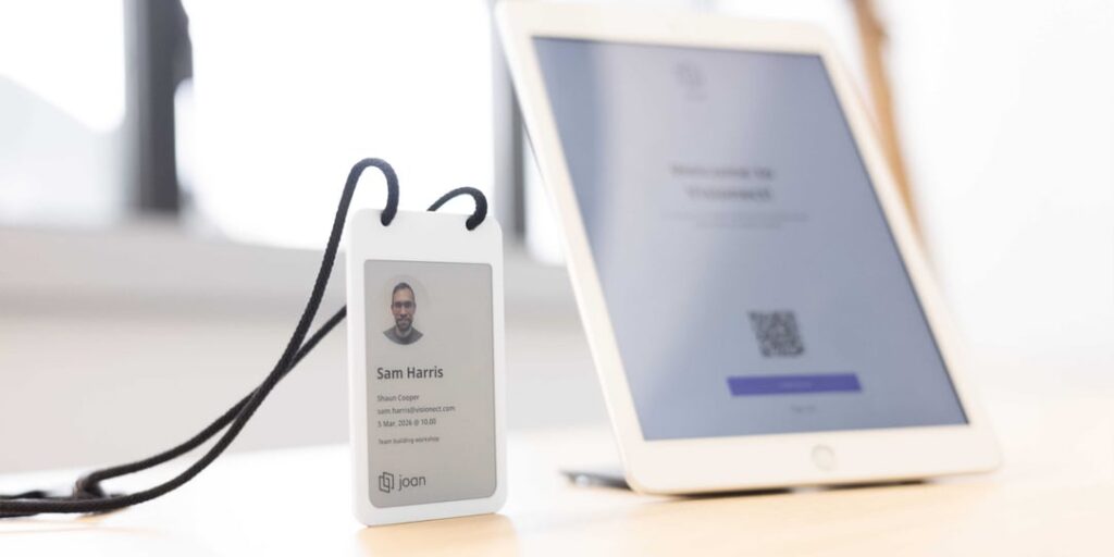 Joan e-paper visitor badge displaying Sam Harris's name, host, and event details, with a check-in tablet in the background.