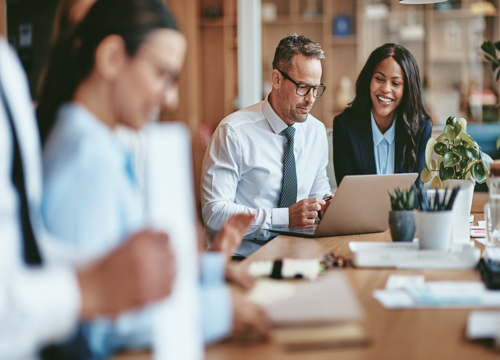 Three business professionals collaborating around a conference table with a laptop in a modern office setting.