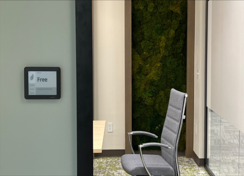 Joan e-ink display mounted outside a modern meeting room showing 'Free' status, with a moss wall and office chair visible inside.