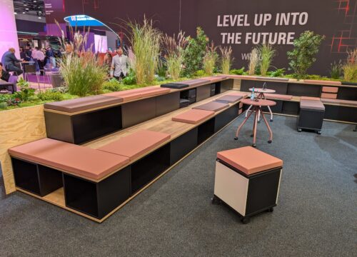 Modular lounge seating with terracotta cushions and storage units at a workplace expo booth with greenery backdrop.