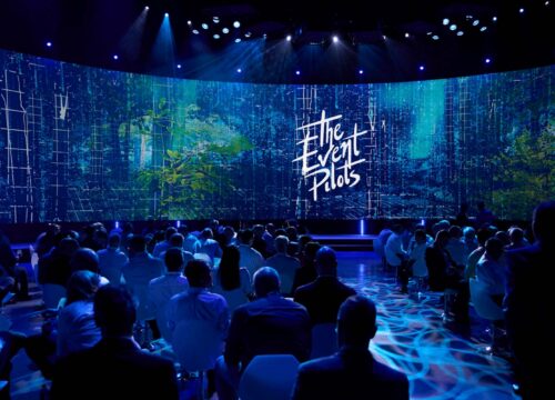 Audience at a large corporate event facing a curved LED display showing 'The Event Pilots' logo amid nature visuals.