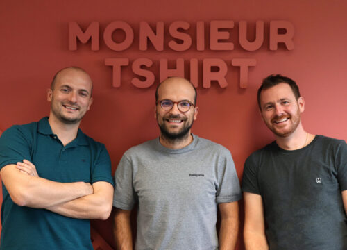 Three smiling men standing together in front of the Monsieur Tshirt branded wall inside their office.