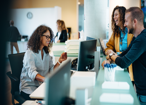 Receptionist smiling and welcoming two visitors at a modern office front desk.