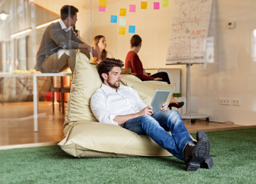 Man working on a tablet in a bean bag chair in a modern activity-based office with colleagues collaborating behind a glass wall.
