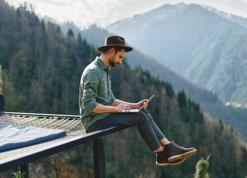 Man in a wide-brimmed hat working on a laptop outdoors on a mountain terrace with scenic forested peaks behind him.
