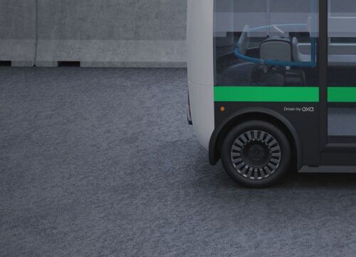 Autonomous electric shuttle bus with a green stripe and 'Driven by OXA' branding parked on a gray surface.