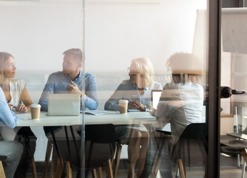 Five professionals collaborating in a glass-walled meeting room with laptops and coffee cups on the table.