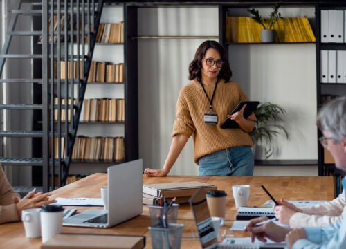 Three professionals in a modern office meeting, one standing and presenting while two colleagues listen and take notes.