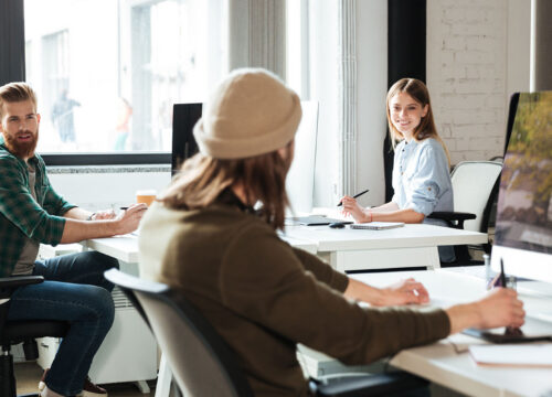 Three colleagues working at desks in a bright open-plan office, smiling and looking aside from their computers.