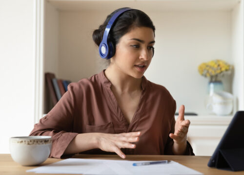 Woman with blue headphones speaking during a video call on a tablet at a home office desk.