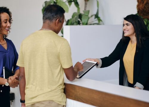 Receptionist handing a tablet to a visitor at a modern office front desk during check-in.