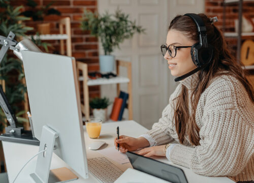 Woman wearing headphones and glasses working at a home office desk with a monitor and graphics tablet.