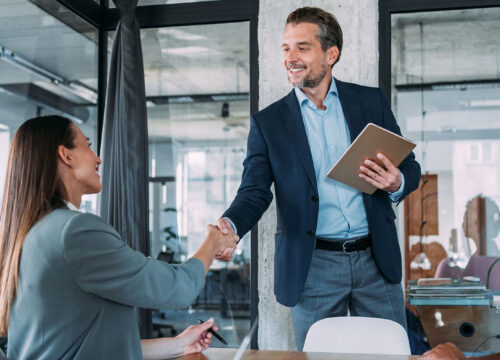 Business professionals shaking hands across a conference table in a modern open-plan office.