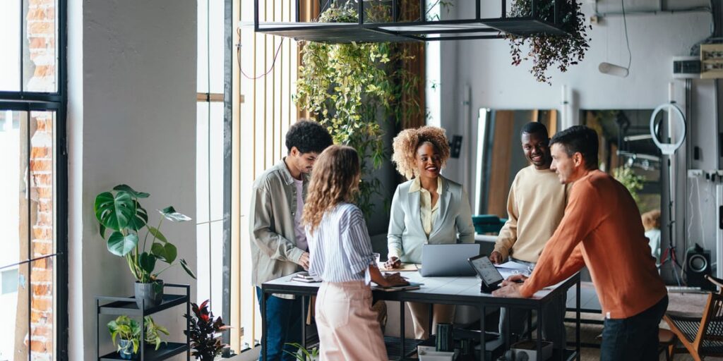 Five diverse professionals collaborating around a standing table in a bright, plant-filled modern office.