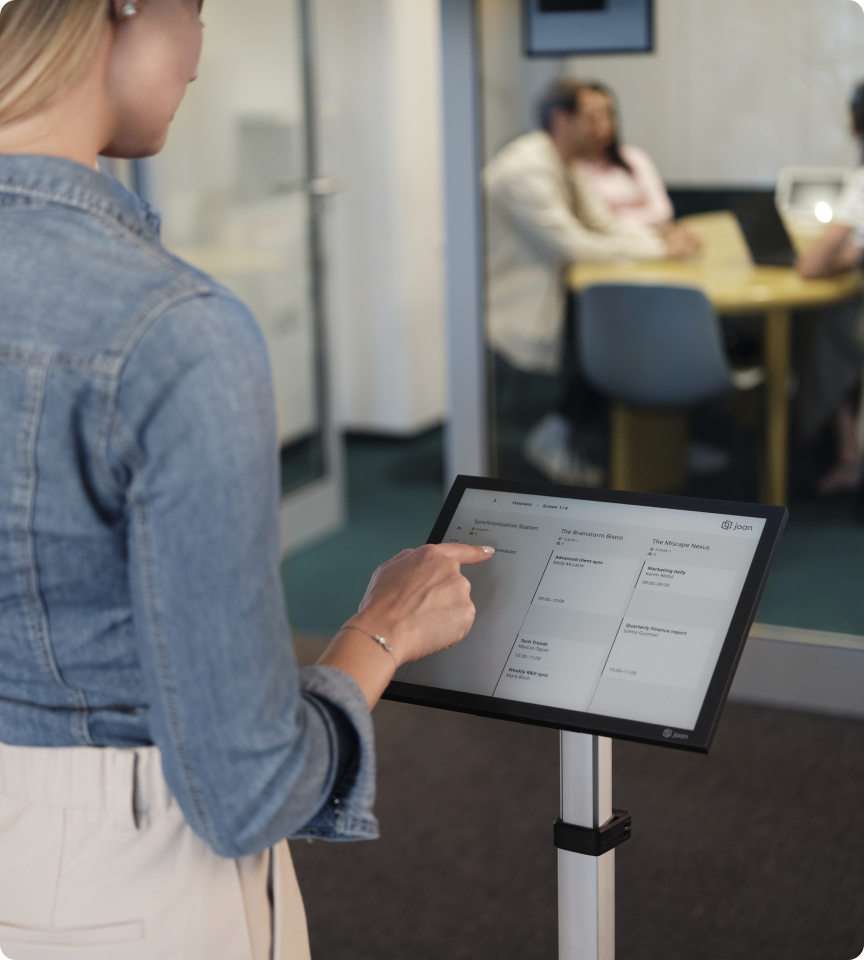 Woman interacting with a Joan 13 e-ink display on a floor stand outside a glass-walled meeting room.