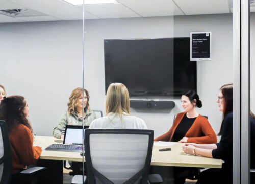 Five women in a meeting around a conference table, with a Joan e-ink room display mounted on the glass wall outside.