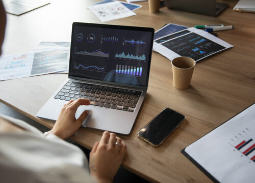 Office worker reviewing financial graphs and dashboards on a laptop surrounded by printed reports and data charts.
