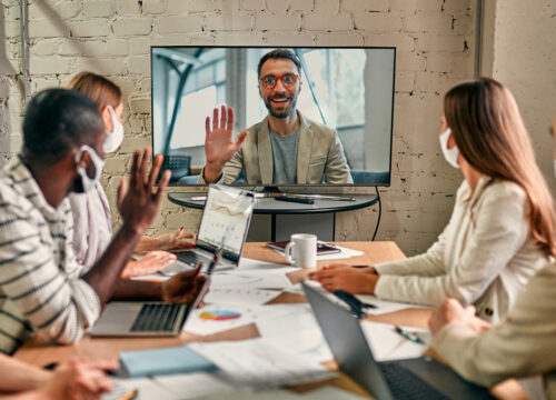Four masked coworkers in a conference room attend a hybrid meeting via video call with a remote colleague on a large screen.