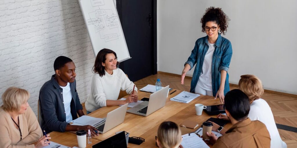 Diverse team of six professionals in a business meeting, with a woman presenting at a wooden conference table.