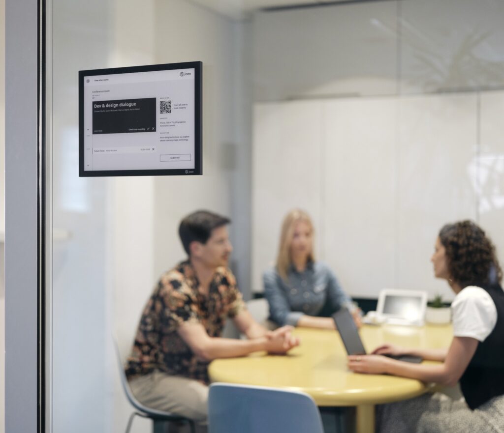 Joan 13 e-ink display mounted on a glass wall showing a conference room booking with three colleagues meeting inside.