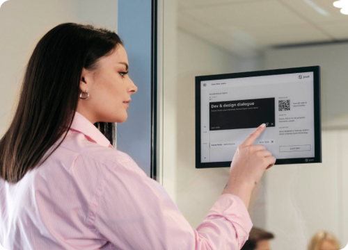 Woman touching a Joan e-ink display mounted on a glass wall showing a room booking schedule and QR code.