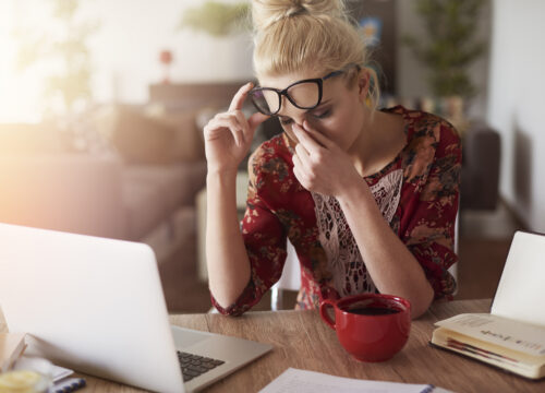 Woman at desk pinching her nose in pain, with laptop, coffee, and notebook nearby.